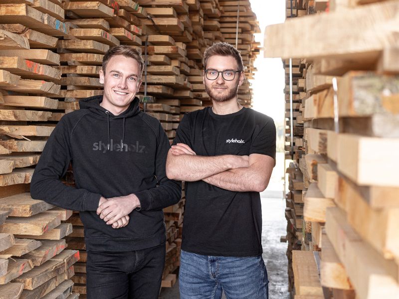 Die Gründer von Styleholz vor meterhohen Stapeln des Rohmaterials, aus dem die Woodsticks hergestellt werden